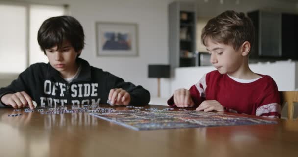 若い男の子の焦点を当てたデュオは 屋内の木のテーブルに活気のあるジグソーパズルのチームワークに従事し 問題解決のスキルを披露しています — ストック動画
