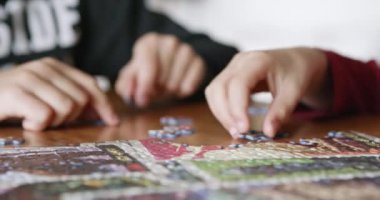 İki çocuk, ahşap bir masada renkli bir yapboz yapmaya odaklanırken, elleri dikkatle parçaları seçiyor..