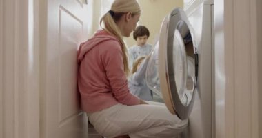 Bir anne, pijama giymiş, çamaşırların çamaşır makinesinde dönüşünü seyreden, ev işleri üzerine bağ kuran çocuklarını denetler..