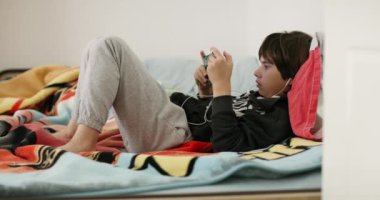 Bir çocuk yatağında rahat bir şekilde uzanır, becerikli elleriyle yönettiği el bilgisayarı oyun konsoluna odaklanır..
