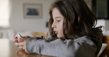 Uzun saçlı bir çocuk akıllı telefonuna dikkatle odaklanırken gülümsüyor, oturma odasındaki masada rahatça oturuyor, muhtemelen bir oyun oynuyor ya da internette geziniyor..