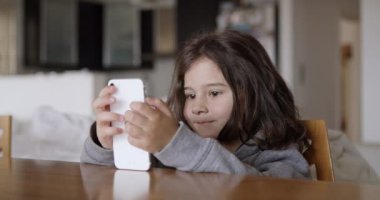 Uzun saçlı bir çocuk akıllı telefonuna dikkatle odaklanırken gülümsüyor, oturma odasındaki masada rahatça oturuyor, muhtemelen bir oyun oynuyor ya da internette geziniyor..