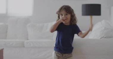 Mavi tişörtlü, uzun kıvırcık saçlı genç bir çocuk gülümsüyor ve arka planda yastıklar ve kanepe ile iyi aydınlatılmış bir oturma odasında el sallıyor..