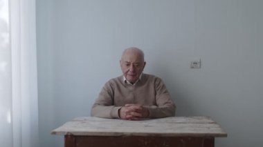 Düşünceli yaşlı bir adam, elleri ahşap bir masanın üzerinde minimuma döşenmiş bir odada derin derin düşünür bir ifadeyle oturur..