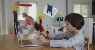 Genç bir çocuk yemek masasındaki dizüstü bilgisayarı üzerinde özenle çalışıyor, aile üyeleri hareketli bir ev ortamında gezerken, gürültü yaparken, oyun oynarken..