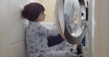 Desenli pijamalı genç bir çocuk giysilerini açık bir çamaşır makinesine dikkatlice yerleştiriyor. Bu da bağımsızlık ve sorumluluk göstergesi..