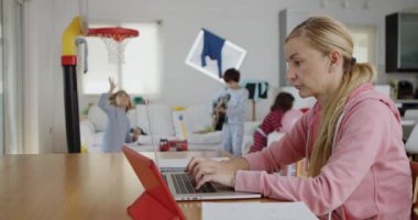 Pembe kazaklı, odaklanmış bir anne oturma odasında dizüstü bilgisayarıyla çalışıyor. Çocukları da etrafı kirletiyor..