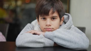 Düşünceli bir ifadeye sahip genç bir çocuk, çenesini çapraz kollarının üzerinde bir masanın üzerinde, gri bir kapüşonlu giymiş ve doğrudan ileriye bakıyor..