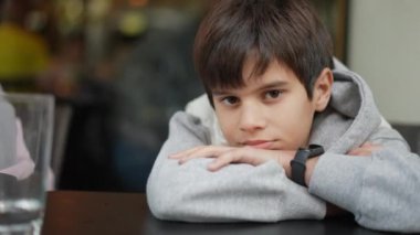 Düşünceli bir ifadeye sahip genç bir çocuk, çenesini çapraz kollarının üzerinde bir masanın üzerinde, gri bir kapüşonlu giymiş ve doğrudan ileriye bakıyor..