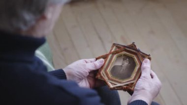 Kıdemli bir kadın, bir askerin eski çerçeveli bir fotoğrafını beşiğinde tutuyor, bir yansıma anı yakalıyor ve hatırlıyor..