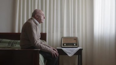Yaşlı bir adam, hafif aydınlatılmış bir odada yatağın kenarında oturuyor. Yüzünde hüzünlü bir ifade, yanında eski moda bir radyo var..