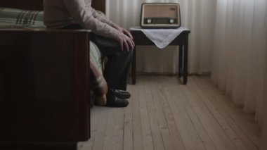 Yaşlı bir adam, hafif aydınlatılmış bir odada yatağın kenarında oturuyor. Yüzünde hüzünlü bir ifade, yanında eski moda bir radyo var..