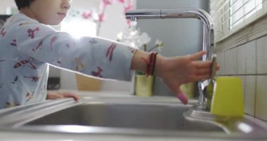 Bir çocuk musluktan su içmek için mutfak lavabosunun üzerine eğiliyor. Gün ışığı manzarayı aydınlatıyor..