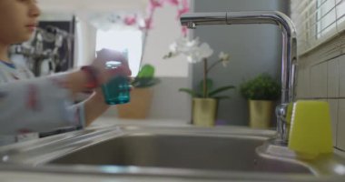 Bir çocuk paslanmaz çelikten bir bardağı dikkatlice doldurur. Lavabo musluğu, doğal gün ışığı. Samimi mutfak sahnesini aydınlatır..