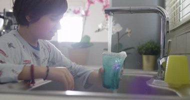 Bir çocuk paslanmaz çelikten bir bardağı dikkatlice doldurur. Lavabo musluğu, doğal gün ışığı. Samimi mutfak sahnesini aydınlatır..