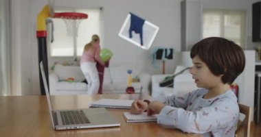 Genç bir çocuk yemek masasındaki dizüstü bilgisayarı üzerinde özenle çalışıyor, aile üyeleri hareketli bir ev ortamında gezerken, gürültü yaparken, oyun oynarken..
