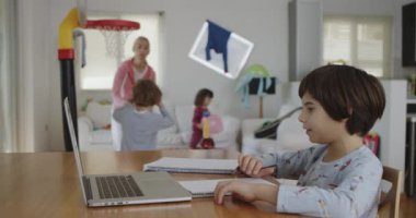 Genç bir çocuk yemek masasındaki dizüstü bilgisayarı üzerinde özenle çalışıyor, aile üyeleri hareketli bir ev ortamında gezerken, gürültü yaparken, oyun oynarken..