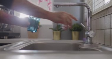 Bir insan, mutfaktaki lavabonun üzerine modern paslanmaz çelik bir musluktan temiz mavi bir bardağa su dökerken görülüyor..