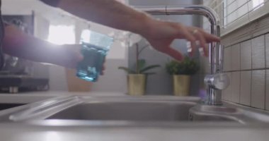 Bir insan, mutfaktaki lavabonun üzerine modern paslanmaz çelik bir musluktan temiz mavi bir bardağa su dökerken görülüyor..
