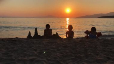 Çocuklar da dahil kumsalda oturan bir grup insan. Güneş arka planda batıyor, sahne üzerinde sıcak bir parıltı yaratıyor..