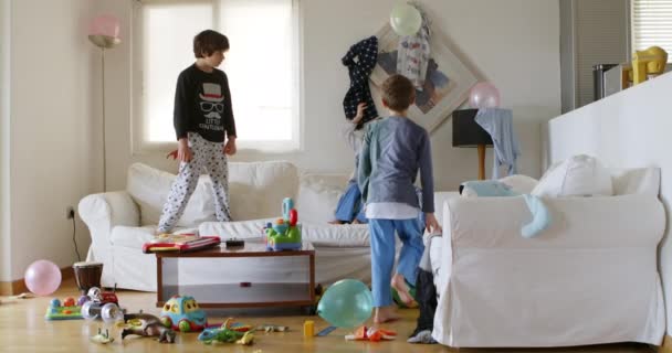 三个孩子正在一间混乱的客厅里精力充沛地跳上一张白色的沙发 沙发上围着玩具和家居用品 阳光从窗户射了进来 — 图库视频影像