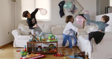 Üç çocuk, dağınık oturma odasındaki oyuncaklar ve ev eşyalarıyla dolu beyaz bir kanepede enerjik bir şekilde zıplıyorlar..