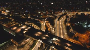 Yoğun bir şehir kavşağı üzerinde gece görüşü, kavis boyunca hareket halindeki araçların parlayan ışıklarını gösteriyor..