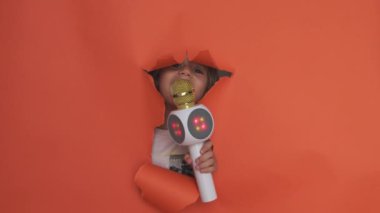 Neşeli bir çocuk elinde parlak bir LED mikrofonla portakal renkli bir arka planda yüzünü açıyor. Eğlenceli bir karaoke ya da şarkı söyleme anı öneriyor..