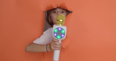 Neşeli bir çocuk elinde parlak bir LED mikrofonla portakal renkli bir arka planda yüzünü açıyor. Eğlenceli bir karaoke ya da şarkı söyleme anı öneriyor..