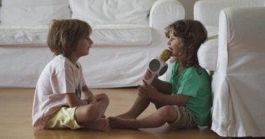 Ahşap zeminde oturan bir çift genç çocuk evlerinin rahatlığında neşeli bir karaoke seansında oyuncak bir mikrofonla büyülü bir müzik anı paylaşıyorlar..
