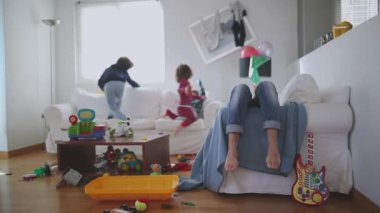 Yorgun anne gözleri kapalı oturur ve kanepeye uzanır. Etrafı oyuncaklarla çevrili, dağınık bir oturma odası, yavaş çekim..