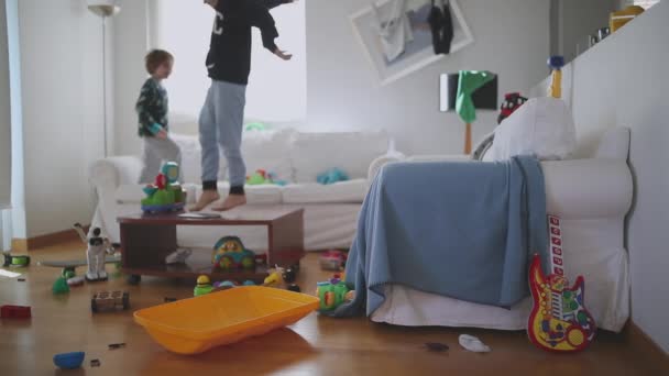 三个孩子正精力充沛地慢吞吞地跳着 在一间杂乱无章的客厅里 躺在一个被玩具和家居用品环绕的白色沙发上 阳光从窗户射进来 — 图库视频影像