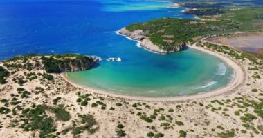Voidokilia Sahili 'nin sakin kıvrımları yukarıdan çekiliyor, turkuaz berrak suların altın kumlu plaja hafifçe dokunduğunu gösteriyor..