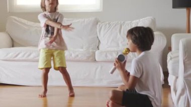 Genç bir çocuk elinde bir karaoke mikrofonuyla yerde oturuyor, coşkuyla şarkı söylüyor, diğer çocuk ise iyi aydınlatılmış bir oturma odasında izliyor..
