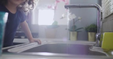 Kıvırcık saçlı tatlı çocuk mutfak lavabosunun üzerine eğilip musluktan su içiyor. Gün ışığı sahneyi aydınlatıyor..