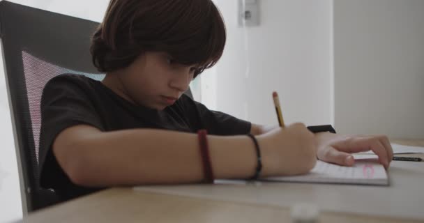 一个小男孩趴在桌子上 全神贯注地做着家庭作业 身边拿着笔和纸 偶尔检查他的腕表 — 图库视频影像