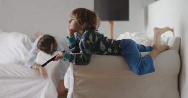 İki çocuk bir oturma odasında aylak aylak yatıyor, biri elinde uzaktan kumandayla kanepede yatıyor, gözleri televizyona yapışık, diğeri yakınlarda oturuyor, uyuşukluk fotoğrafı..