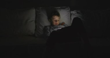 Genç bir çocuk beyaz bir kanepede rahatça oturur. Dikkatini, sakin alacakaranlık saatlerinde elinde tuttuğu kırmızı tablet çeker..
