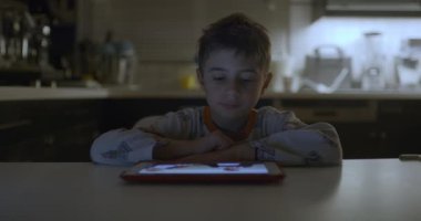 Genç bir çocuk, mutfak masasında çenesini dinlendirirken dijital tablete dikkatle odaklanır. Aygıtın loş ışıklı bir odada ışıltısıyla aydınlanır..