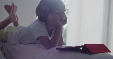 Beyaz bir kanepeye uzanmış bir çocuk, dijital tabletine dalmış, öğle vakti güneşli, modern bir oturma odasında..