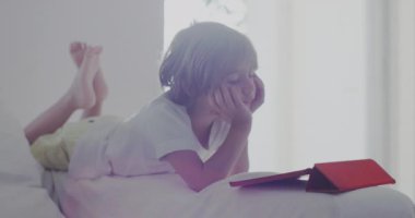 Beyaz bir kanepeye uzanmış bir çocuk, dijital tabletine dalmış, öğle vakti güneşli, modern bir oturma odasında..