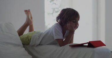 Beyaz bir kanepeye uzanmış bir çocuk, dijital tabletine dalmış, öğle vakti güneşli, modern bir oturma odasında..