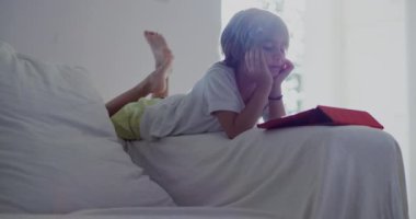 Beyaz bir kanepeye uzanmış bir çocuk, dijital tabletine dalmış, öğle vakti güneşli, modern bir oturma odasında..