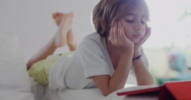 Beyaz bir kanepeye uzanmış bir çocuk, dijital tabletine dalmış, öğle vakti güneşli, modern bir oturma odasında..