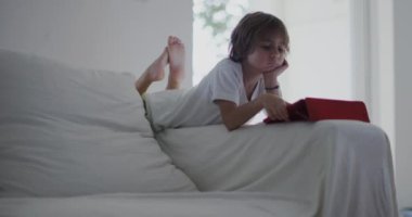 Beyaz bir kanepeye uzanmış bir çocuk, dijital tabletine dalmış, öğle vakti güneşli, modern bir oturma odasında..