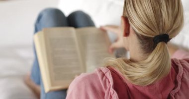 Sarışın, at kuyruklu bir kadın, beyaz bir kanepede rahat bir şekilde uzanırken bir kitaba dalmış, rahat bir hayat sürüyor..