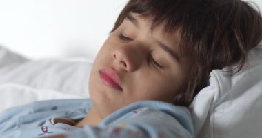 Genç bir çocuk başını beyaz bir yastığa koyar, ciddi bir ifadeyle kafasını çevirir, düşüncelere dalmış gibi görünür..