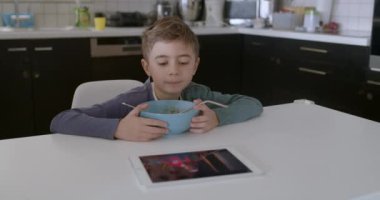 Genç bir çocuk modern bir mutfak masasında oturuyor, önünde bir kase mısır gevreğiyle dijital tablete odaklanmış..