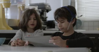 Ağız telleri olan bir çocuk mutfak masasında otururken dijital tabletine dalmış, yanında kahvaltı yapan başka bir çocuk gibi..