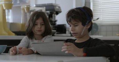 Ağız telleri olan bir çocuk mutfak masasında otururken dijital tabletine dalmış, yanında kahvaltı yapan başka bir çocuk gibi..